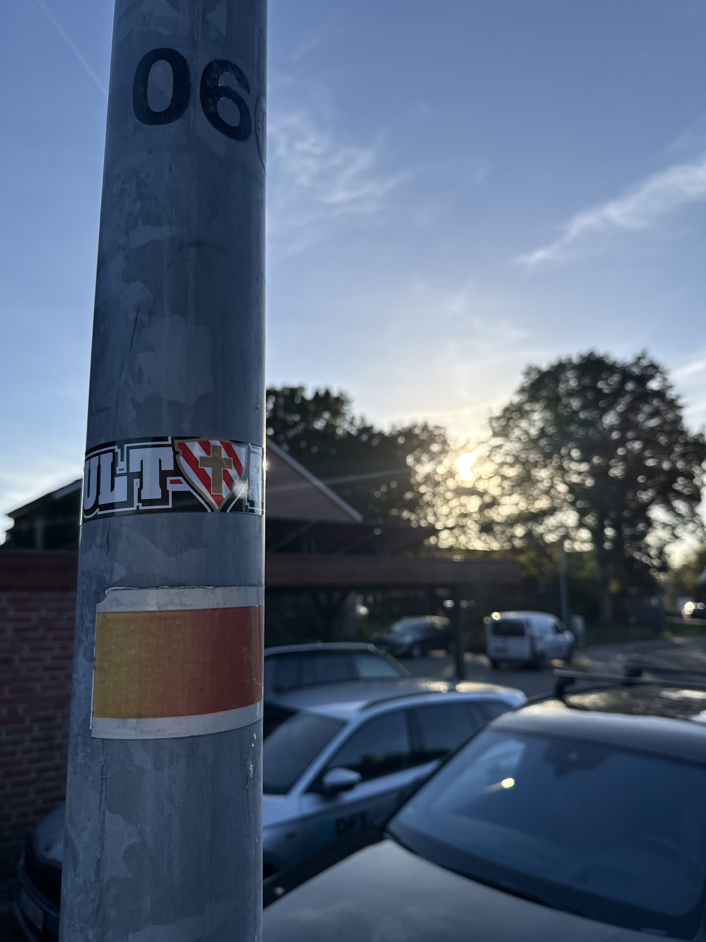 A rectangular sticker wrapped around a grey metal utility pole. The sticker features the bold white text 'ULT' with a black outline on the left, followed by a central shield emblem. The shield is divided diagonally into red and white sections and features a gold cross in the center. To the right of the shield, the sticker continues around the pole. Below it, a second, simpler sticker with a yellow-to-red gradient is also visible.