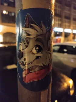 A sticker featuring a stylized cartoon wolf, wearing a red scarf. The wolf has large eyes and a playful expression. The sticker is on a dark blue background, with the artist's website partially visible near the bottom. 