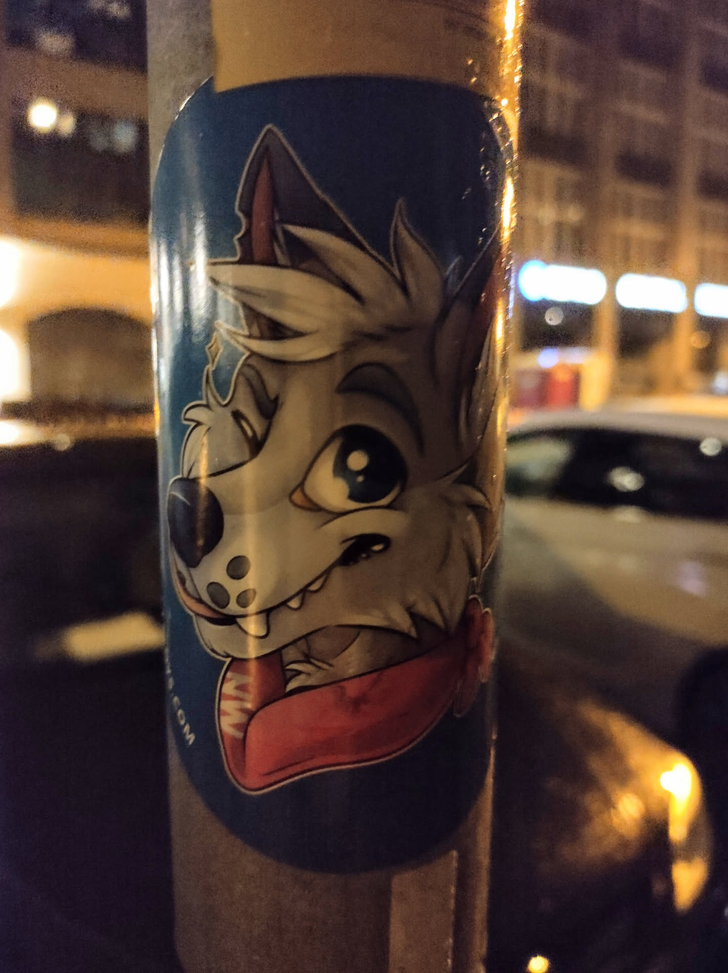 A sticker featuring a stylized cartoon wolf, wearing a red scarf. The wolf has large eyes and a playful expression. The sticker is on a dark blue background, with the artist's website partially visible near the bottom. 
