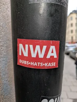 This is a red sticker with the white letters "NWA" on top. Below the letters there are three words separated by dots. Those words are "DUBS", "HATS", and "KASE".