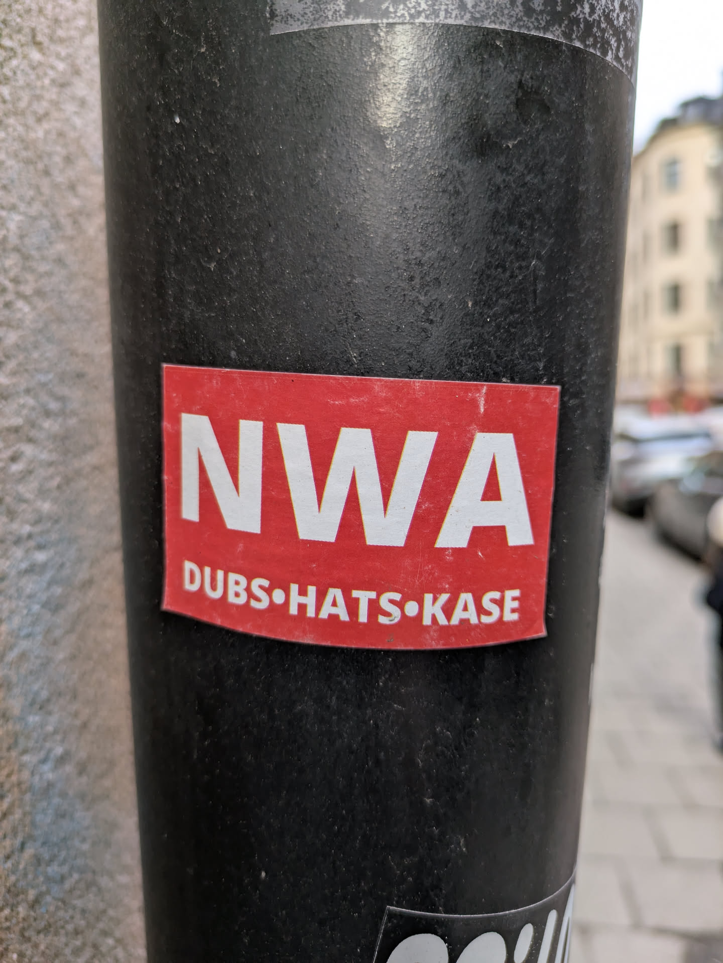 This is a red sticker with the white letters "NWA" on top. Below the letters there are three words separated by dots. Those words are "DUBS", "HATS", and "KASE".