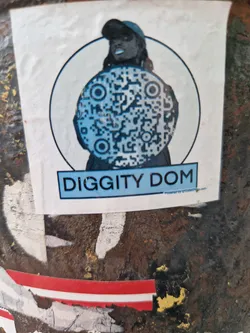 A sticker featuring a stylized illustration of a person holding a large circular object with a QR code. The text 'DIGGITY DOM' is prominently displayed below the illustration.