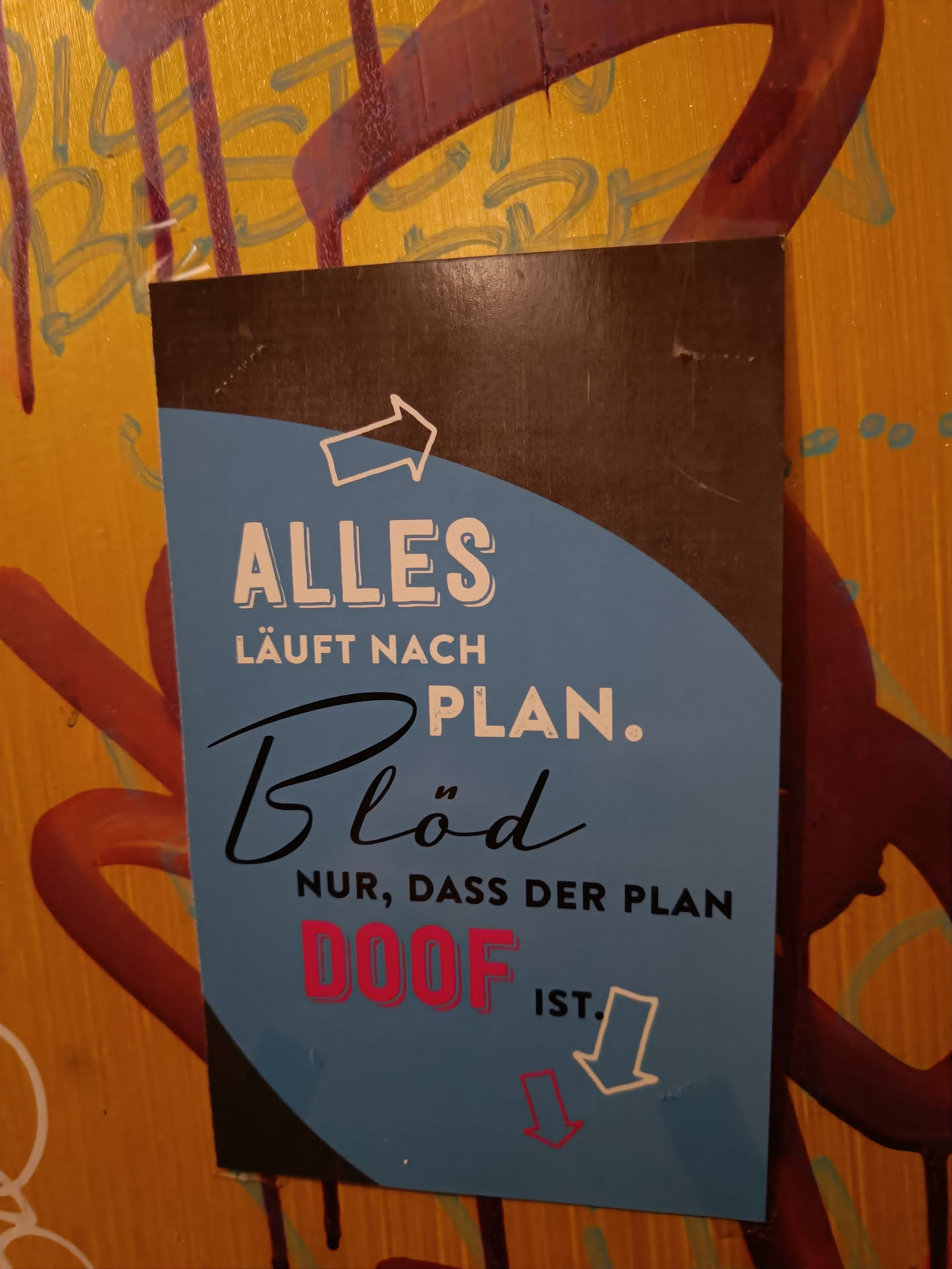 A sticker with a dark brown border and a light blue center. It has a message in German that says: "Everything goes according to plan. Stupidly, only that the plan is stupid."  There are arrows pointing up and down.
