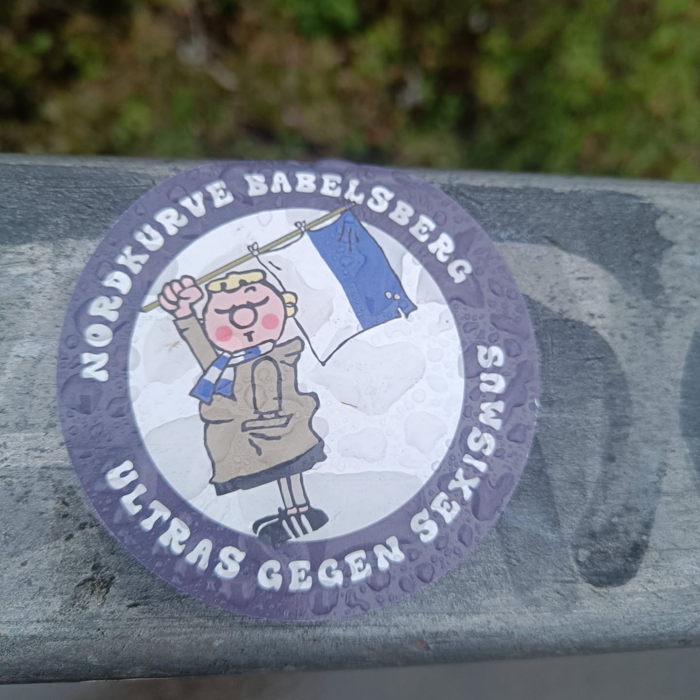 A round sticker with a cartoon figure holding a flag. The text around the figure says "Nordkurve Babelsberg Ultras gegen Sexismus" which translates to "Nordkurve Babelsberg Ultras against Sexism". The sticker appears to be affixed to a railing outdoors.