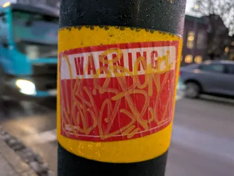 A red sticker with the word WARNING on it.  Someone has tagged over it with yellow graffiti. The sticker is affixed to a dark pole. 