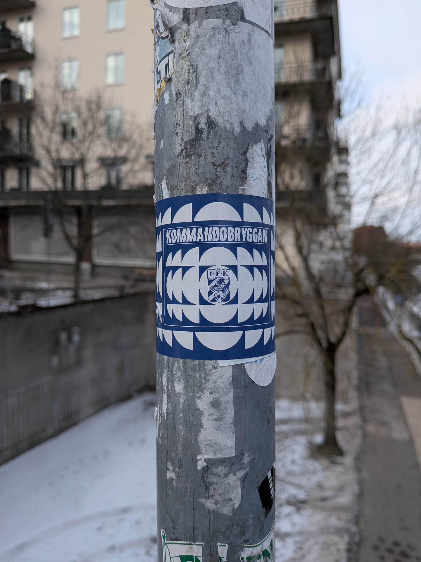 The sticker is rectangular, wrapped around a metal pole, and features a striking design in dark blue and white. At the top, the word "KOMMANDOBRYGGAN" is written in white capital letters against a blue background. The main body of the sticker showcases a repeating geometric pattern composed of white semi-circles and quarter-circles on a blue background, creating a tessellated, almost wave-like or mosaic effect. Centered within this pattern is a white square containing a blue crest or emblem. The crest features a stylized animal (possibly a lion or griffin) and the letters "I.F.K." above it. The overall appearance is clean, graphic, and has a strong visual identity.