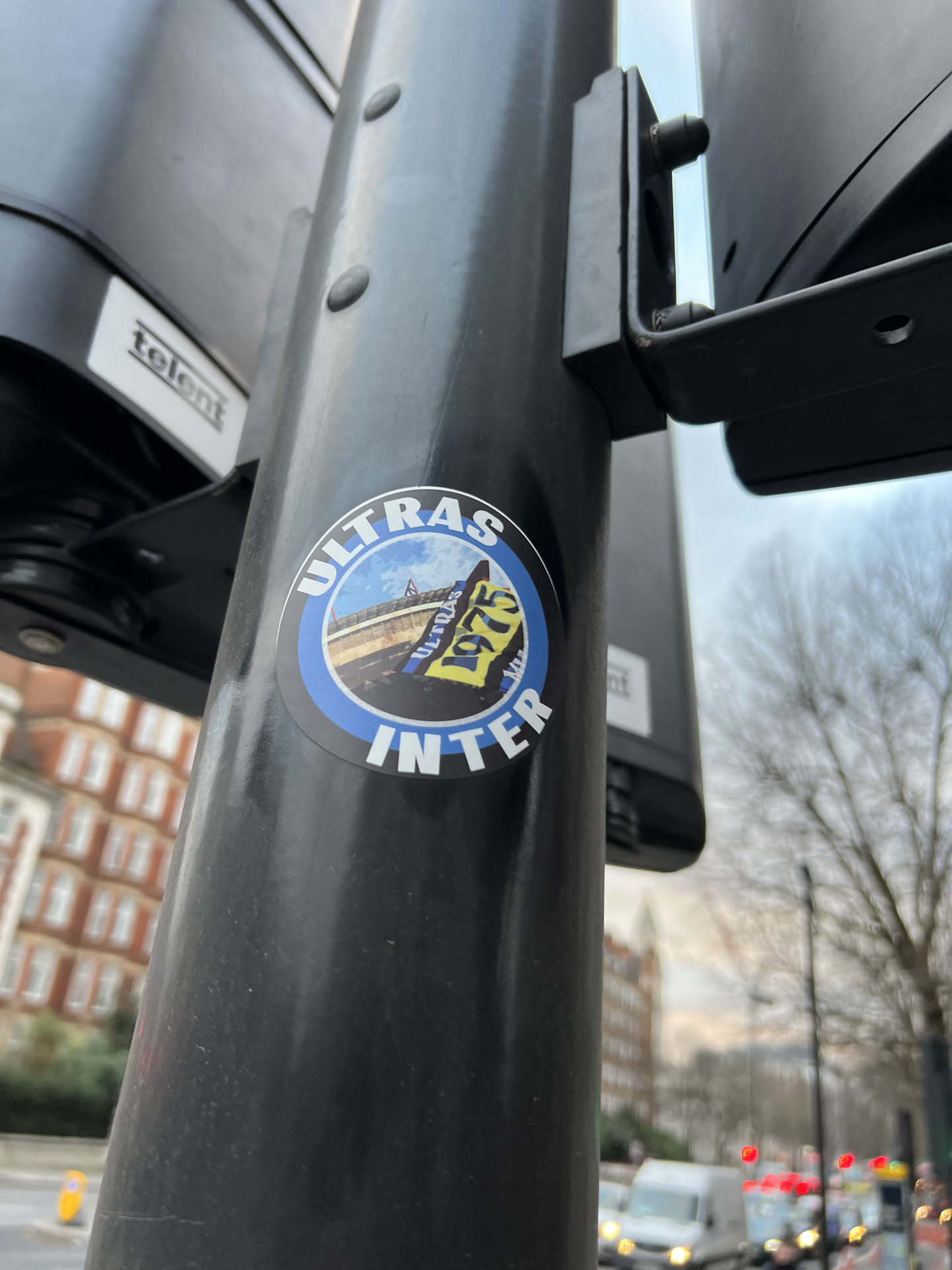 A circular sticker featuring a black outer border and a blue inner border. The central image depicts a football stadium under a blue sky, with several flags or banners in the foreground. A prominent black and yellow/gold flag displays '1975' in white text. White capital letters spell 'ULTRAS' along the top curve and 'INTER' along the bottom curve of the blue border. The overall color scheme includes black, blue, white, and yellow/gold.