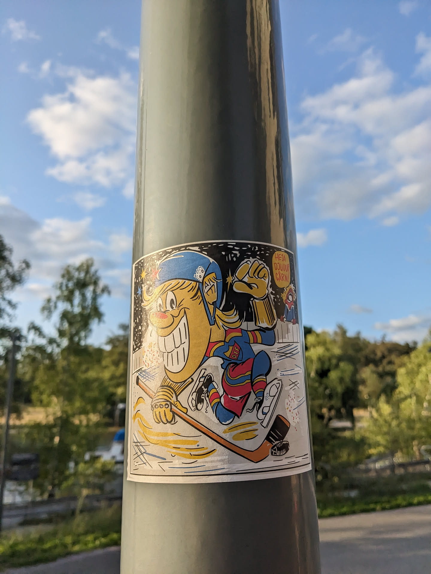 This sticker features a cartoon-like illustration of a hockey player, seemingly happy and enjoying the sport. The style is reminiscent of retro or vintage designs. The background is a simple space-themed design, suggesting a playful and unique approach to the artwork. The sticker is affixed to a gray pole outdoors, suggesting it might be street art.
