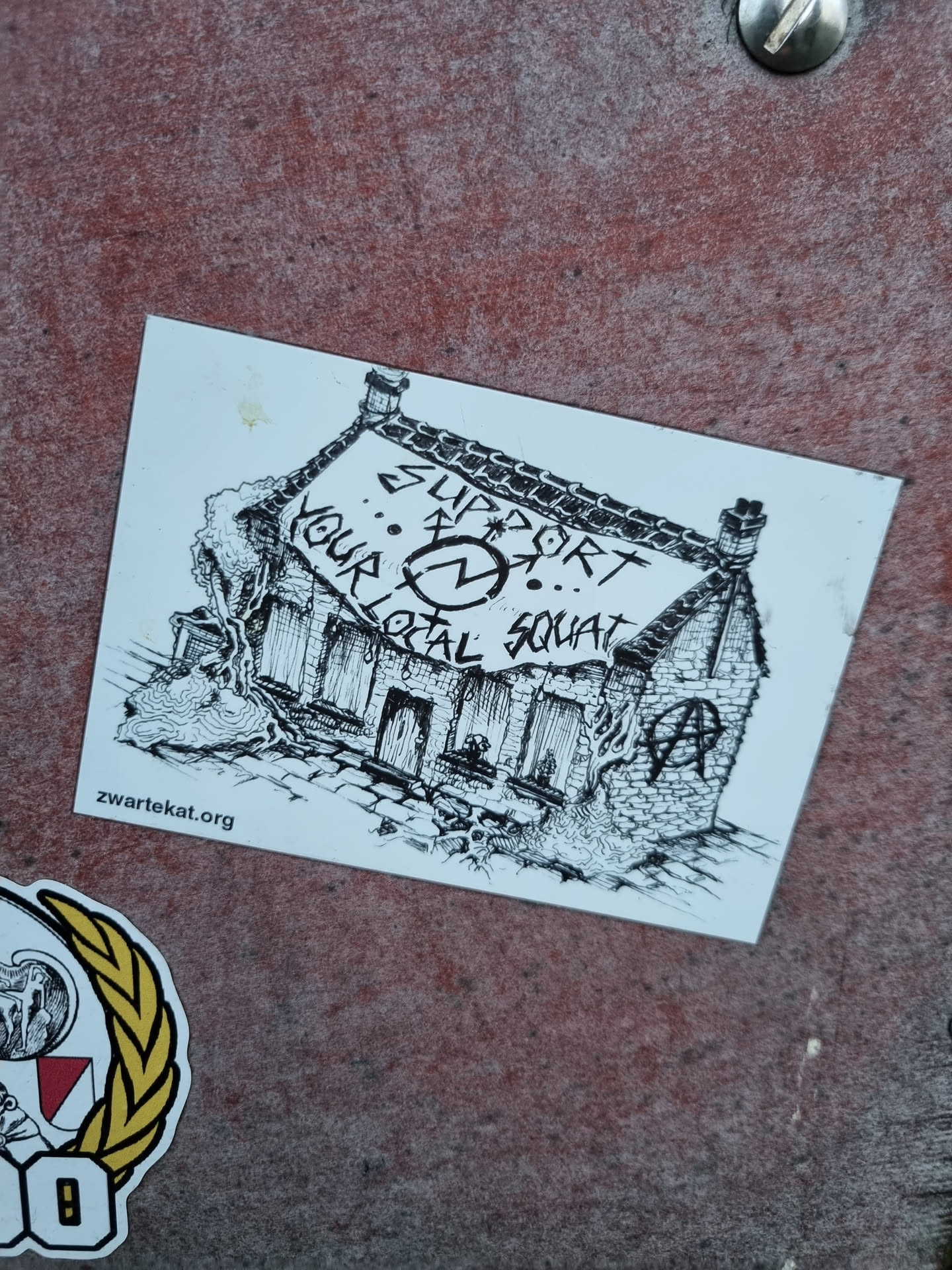 Black and white drawing of a house with a banner that says "Support your local squat".  There is an anarchy symbol on the house. The website zwartekat.org is printed at the bottom.