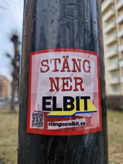 A sticker with the text "Stång ner Elbit" (Shut down Elbit) and a QR code leading to stångnereelbit.se. The sticker is red and yellow, and it's pasted on a black pole.