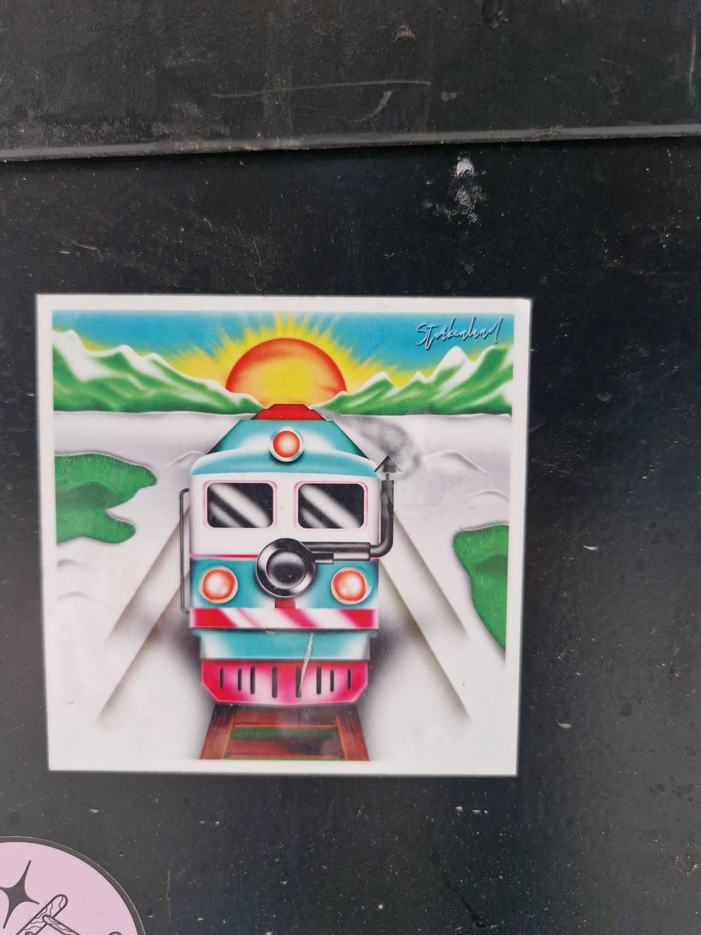 A sticker featuring a stylized illustration of a train approaching the viewer against the backdrop of a sunrise over mountains. The art style is reminiscent of retro or vintage designs.  The train is teal, white, and pink.