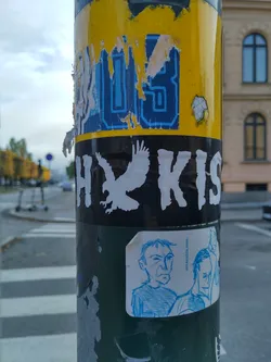 A sticker featuring a stylized eagle and the text HYKIS.  Below it is a smaller sticker with a hand-drawn sketch of two men. The main sticker has a distressed look, perhaps suggesting age or intentional wear.