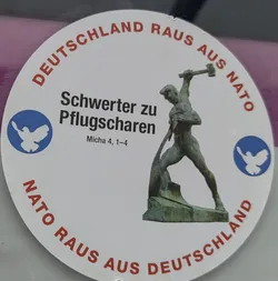 A circular white sticker with a central image of a bronze-colored statue depicting a muscular man forging a sword into a plowshare with a hammer. This iconic 'Swords into Plowshares' sculpture is flanked by two blue circles, each containing a white stylized peace dove. Curved around the top edge of the sticker, in red uppercase letters, is the text 'DEUTSCHLAND RAUS AUS NATO' (Germany out of NATO). Similarly, curved around the bottom edge, also in red uppercase letters, is 'NATO RAUS AUS DEUTSCHLAND' (NATO out of Germany). In the center-left, in bold black text, it reads 'Schwerter zu Pflugscharen' (Swords into Plowshares), with a smaller biblical reference 'Micha 4, 1–4' below it.