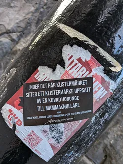 The sticker is black with white text, and is placed on top of a larger, partially torn sticker that has a red and white checkered pattern with red graphic elements. The main sticker contains a message in Swedish and English. At the bottom left, there is a QR code, and a signature is present at the bottom right.