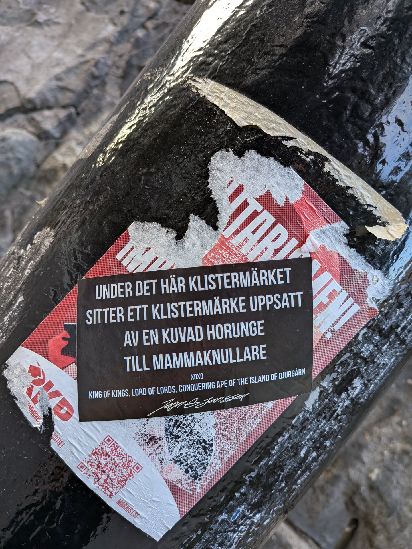 The sticker is black with white text, and is placed on top of a larger, partially torn sticker that has a red and white checkered pattern with red graphic elements. The main sticker contains a message in Swedish and English. At the bottom left, there is a QR code, and a signature is present at the bottom right.