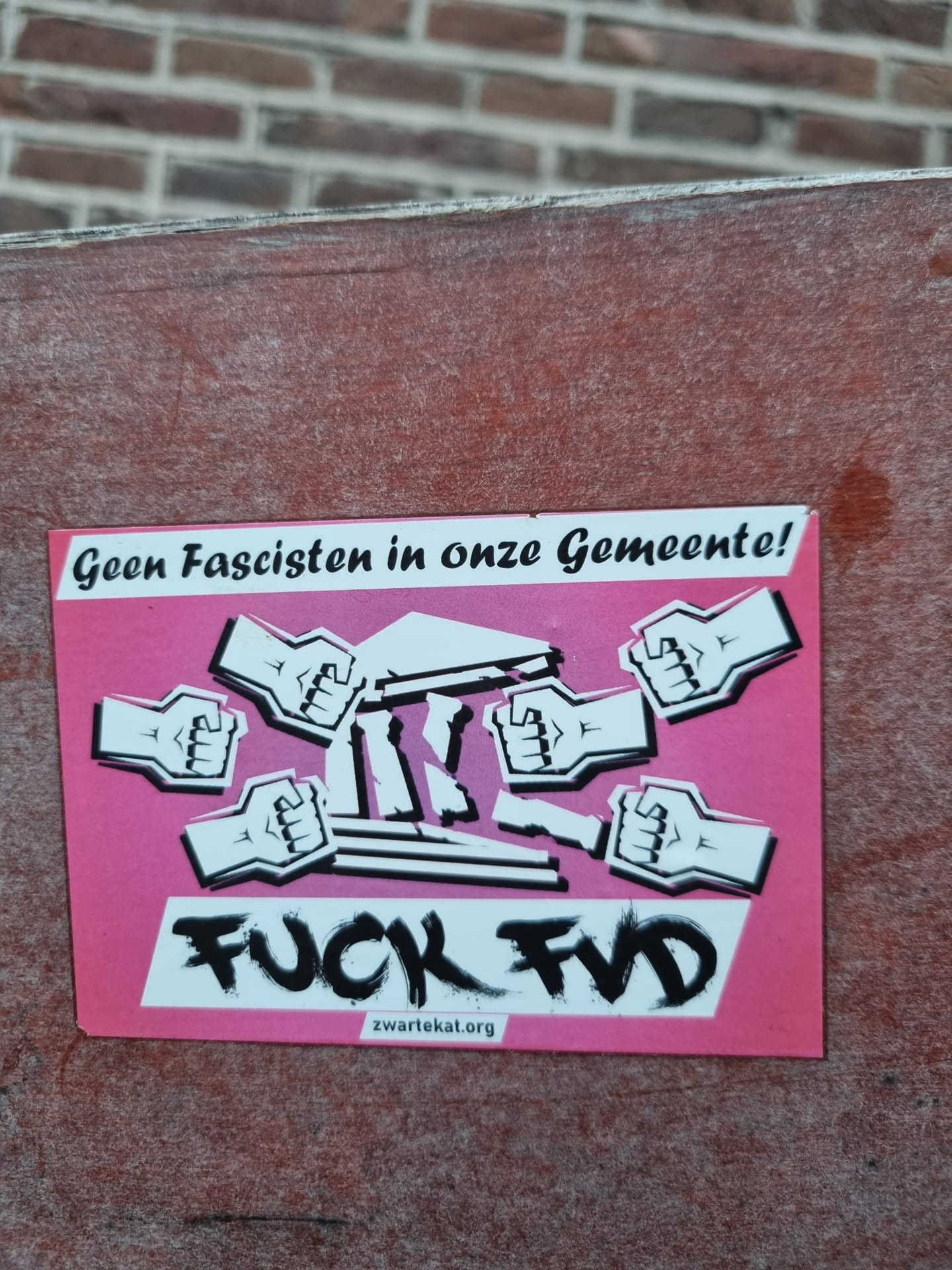 Pink sticker with a illustration of several fists smashing a column. The text on the sticker says in Dutch: 'Geen Fascisten in onze Gemeente!' and 'FUCK FVD'.  There is also a website address at the bottom.