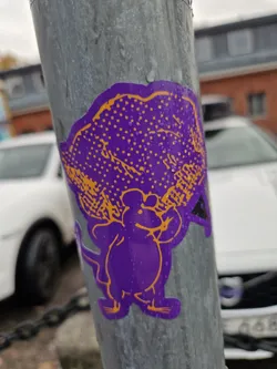 A purple mouse sticker with an orange cloud-like object on its head. The sticker is affixed to a gray metal pole. The background shows part of a parking lot and some buildings.