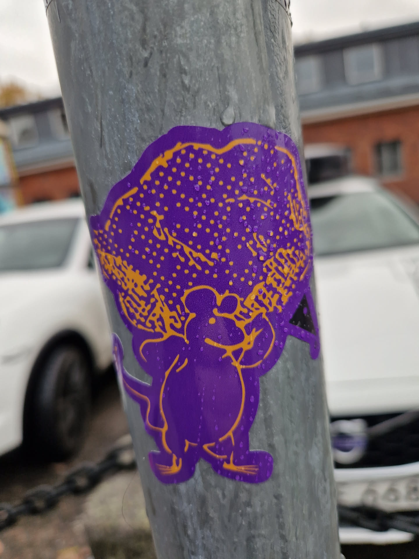 A purple mouse sticker with an orange cloud-like object on its head. The sticker is affixed to a gray metal pole. The background shows part of a parking lot and some buildings.