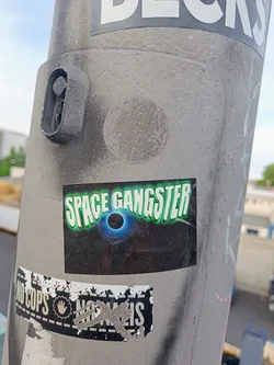 A sticker featuring the text "Space Gangster" in a stylized green font. The sticker has a dark background with a depiction of a black hole or similar celestial phenomenon.