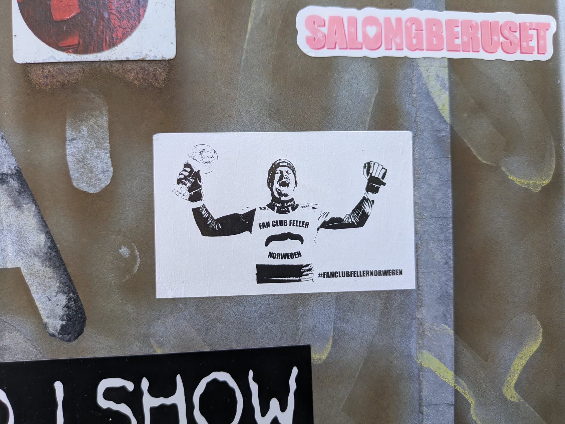 The sticker features a black and white graphic of a male athlete, possibly a skier, with a helmet and sporting gear, raising his arms in triumph. He holds a trophy in his left hand and a clenched fist in his right. Below his image, the text 'FAN CLUB FELLER' is visible, followed by a stylized mustache graphic, and then 'NORWEGEN'. At the very bottom, a hashtag '#FANCLUBFELLERNORWEGEN' is displayed.