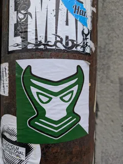 A sticker featuring a stylized mask with horns, primarily green and white. The mask has a bold outline and angular features, giving it a somewhat sinister appearance. The sticker is affixed to a rusty metal pole, surrounded by other stickers. 