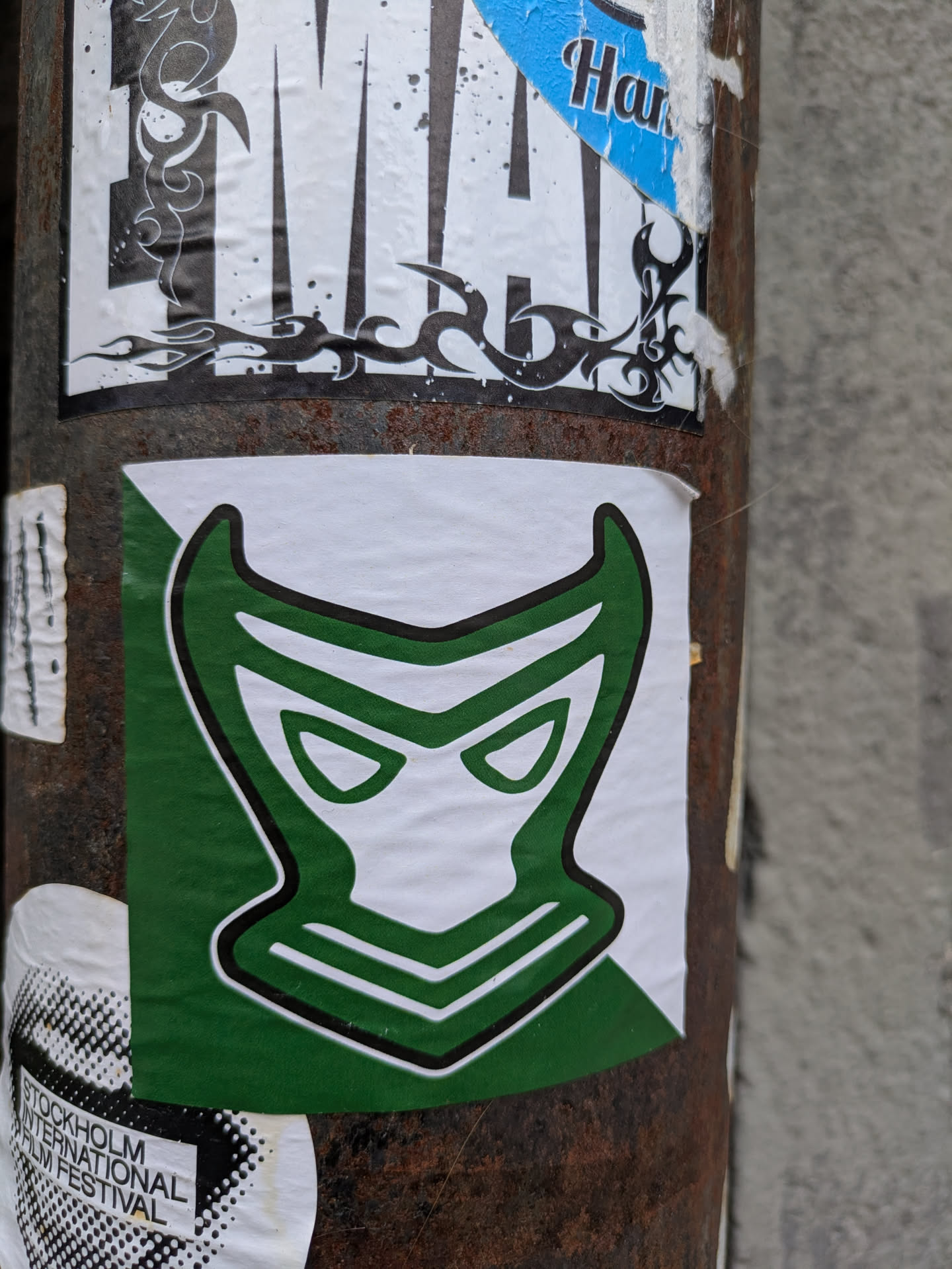 A sticker featuring a stylized mask with horns, primarily green and white. The mask has a bold outline and angular features, giving it a somewhat sinister appearance. The sticker is affixed to a rusty metal pole, surrounded by other stickers. 
