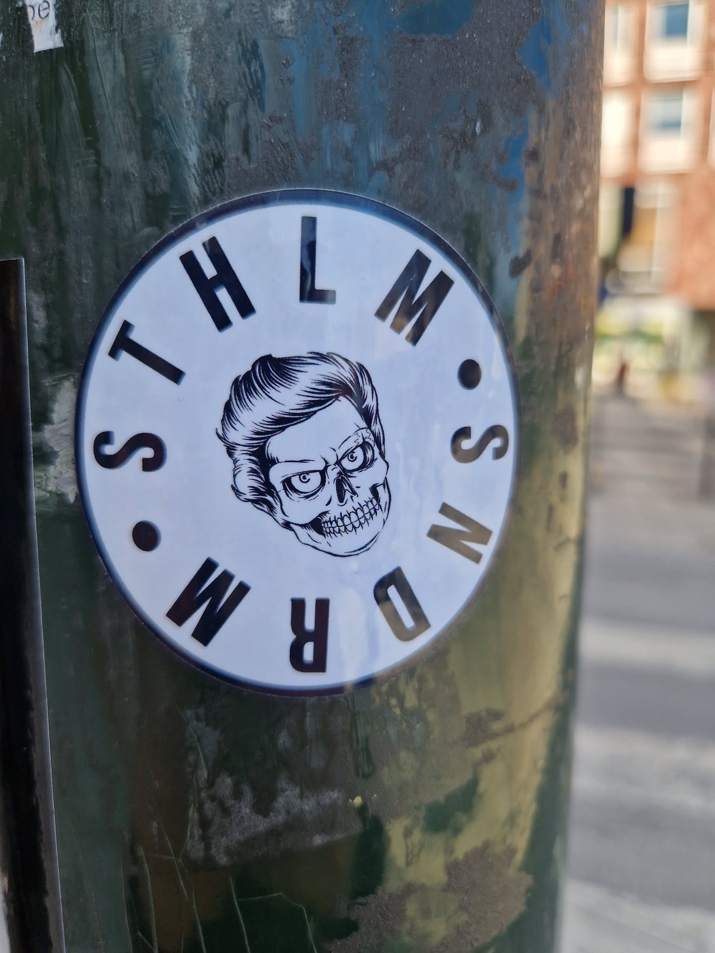 A round sticker featuring a skull with slicked-back hair inside a circle with the letters  'STHLMSNDRM' arranged around the perimeter. The style is black and white with a bold graphic look. 
