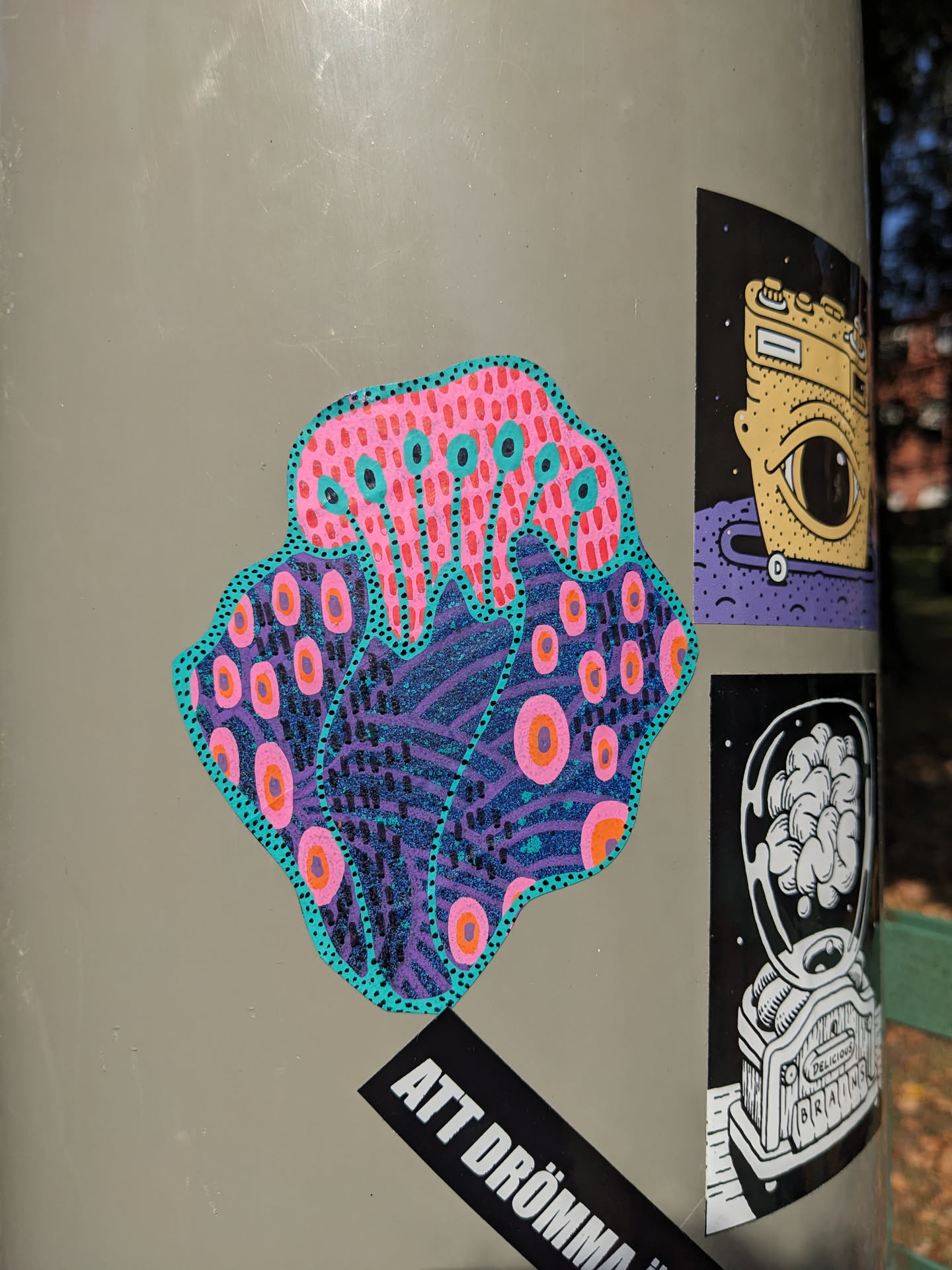 Close-up view of a vibrant sticker featuring a stylized coral or sea anemone with pink, purple, teal, and orange patterns. The artwork is detailed and intricate, with a psychedelic feel.  It's adhered to a grey surface alongside other stickers.