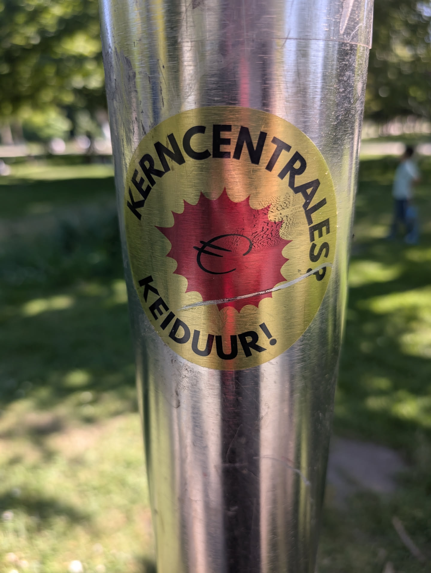 This sticker on a pole has a golden background with a red burst shape in the center, superimposed with a euro currency symbol. The text "KERNCENTRALES? KEIDUUR!" is printed around the edge.