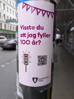 A sticker posted on a pole, promoting Stockholm City's 100th anniversary. The sticker features purple garland at the top, followed by text asking if you knew "I am turning 100 years old?". Below this is an illustration of a traffic light surrounded by stars, a QR code, and the Stockholm City logo.