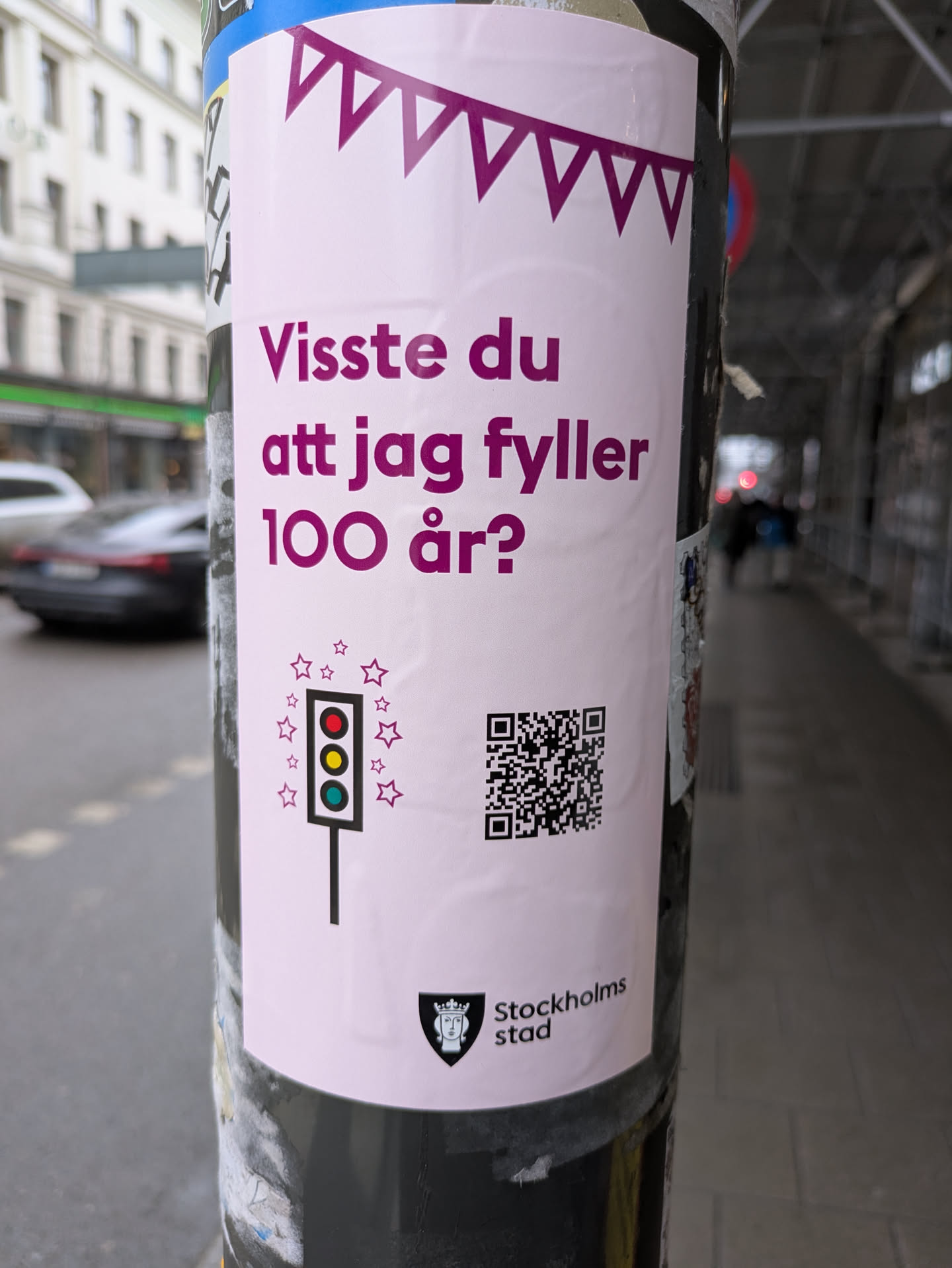 A sticker posted on a pole, promoting Stockholm City's 100th anniversary. The sticker features purple garland at the top, followed by text asking if you knew "I am turning 100 years old?". Below this is an illustration of a traffic light surrounded by stars, a QR code, and the Stockholm City logo.