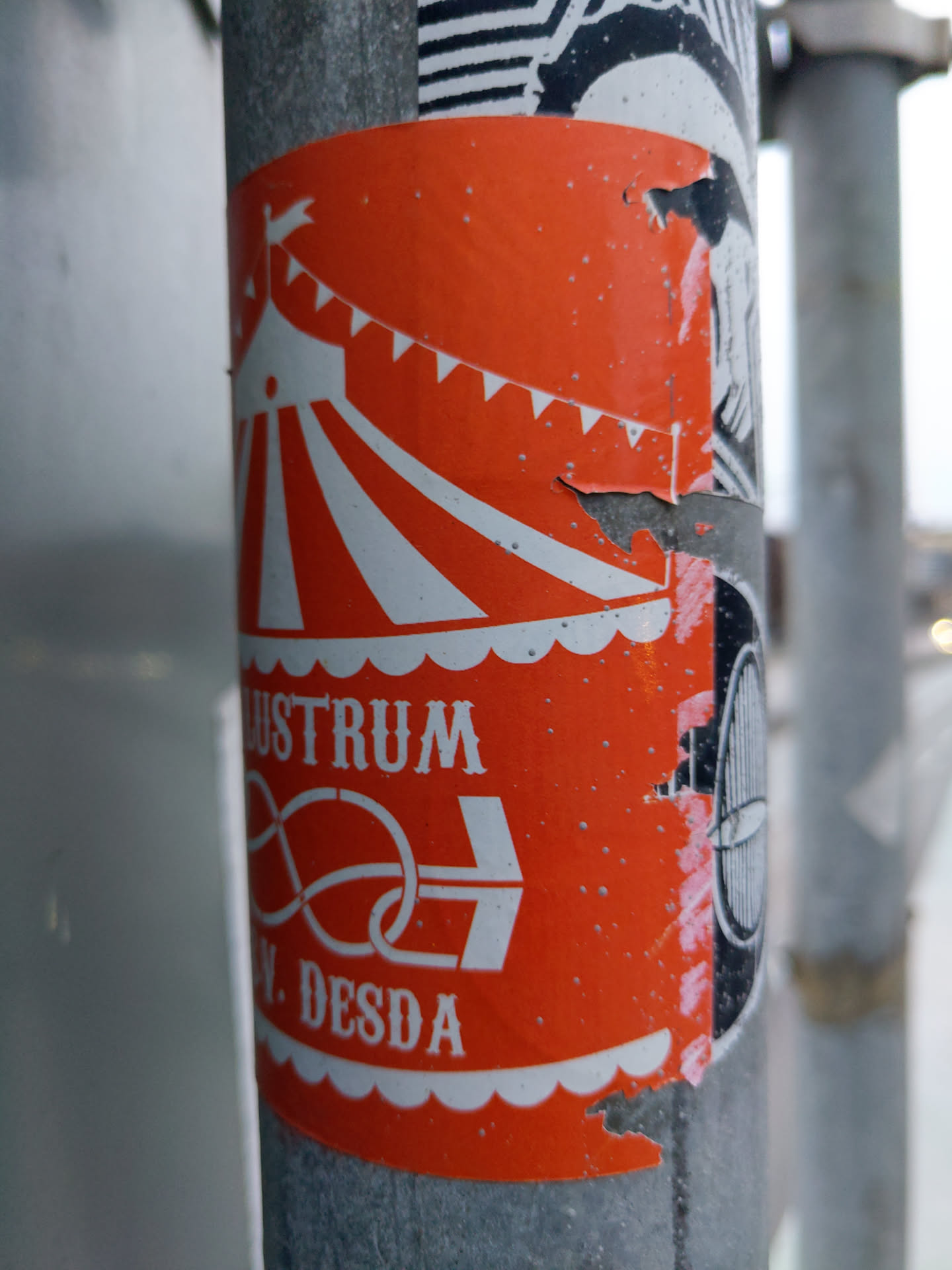 An orange sticker featuring a simple illustration of a circus tent with the word 'lustrum' and a symbol below it.  The sticker shows some wear and tear, suggesting it has been outdoors for a while. The bottom features the text 'N. DESDA'.