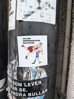 The sticker features a cartoonish illustration on a white background. A large, muscular man, resembling a strongman or wrestler, is depicted in a dark red singlet with matching red boots. He has a dark mustache, dark hair, and visible tattoos on his left bicep and right forearm. He is leaning back, seemingly being lifted or supported by a much smaller child. The child wears a blue short-sleeved shirt, green shorts, yellow and black striped socks, and black shoes. The child has orange hair and is reaching up with both hands to support the strongman's left arm. The overall style is simple and illustrative with clear black outlines.