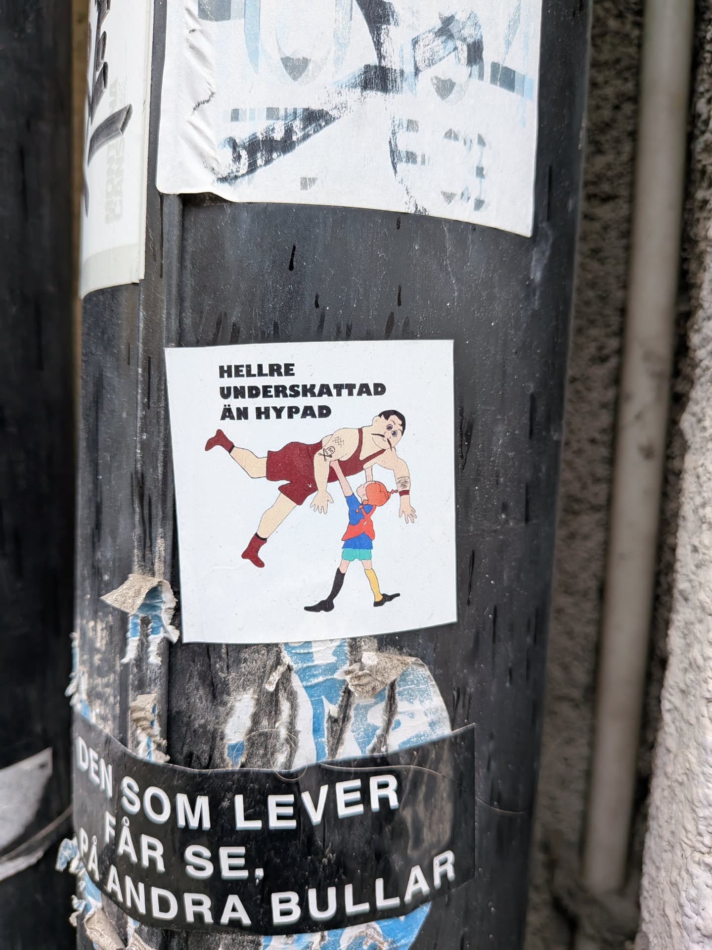 The sticker features a cartoonish illustration on a white background. A large, muscular man, resembling a strongman or wrestler, is depicted in a dark red singlet with matching red boots. He has a dark mustache, dark hair, and visible tattoos on his left bicep and right forearm. He is leaning back, seemingly being lifted or supported by a much smaller child. The child wears a blue short-sleeved shirt, green shorts, yellow and black striped socks, and black shoes. The child has orange hair and is reaching up with both hands to support the strongman's left arm. The overall style is simple and illustrative with clear black outlines.