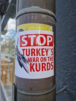 A sticker expressing protest against Turkey's actions against the Kurds. It features an image of a fighter jet and the text 'STOP TURKEY'S WAR ON THE KURDS' in bold red letters.