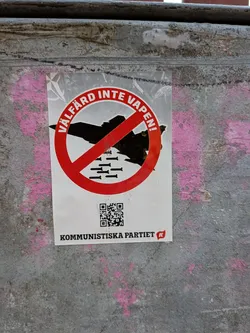 The sticker features a prominent red circle with a diagonal line through it, indicating prohibition. Inside the circle, a silhouette of a jet fighter is depicted, with several bombs falling from it. The text 'VÄLFÄRD INTE VAPEN!' is written in white capital letters along the curve of the red prohibition symbol. Below this, a QR code is visible, and further down, the text 'KOMMUNISTISKA PARTIET' is printed in black, accompanied by a small red logo with a white 'K'. The sticker is white with red and black elements, and the background of the sticker appears to be a light grey with a faint NATO symbol visible in the upper left corner.