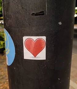 A small square sticker with a red heart on it. It is placed on a dark grey pole.