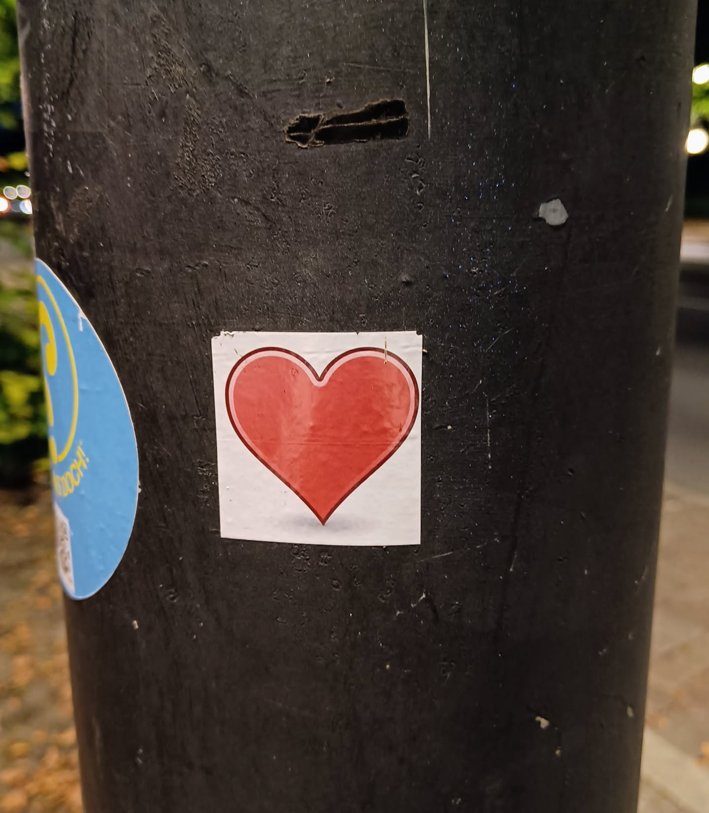 A small square sticker with a red heart on it. It is placed on a dark grey pole.