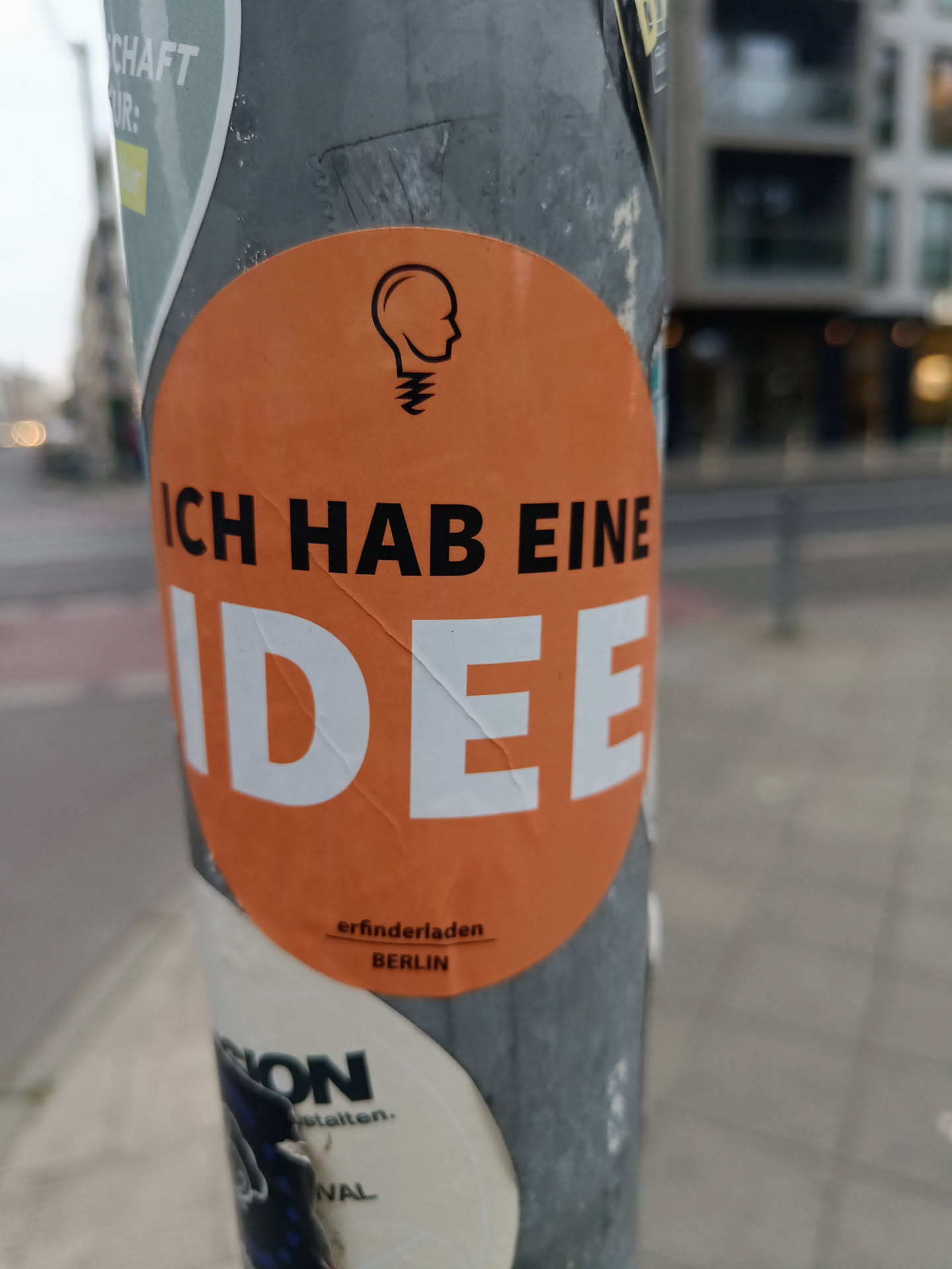 An orange round sticker with a lightbulb head graphic and the text "ICH HAB EINE IDEE" (I have an idea) in bold black letters.  At the bottom, it says "erfinderladen BERLIN" in a smaller font. The sticker is affixed to a gray pole amongst other stickers.