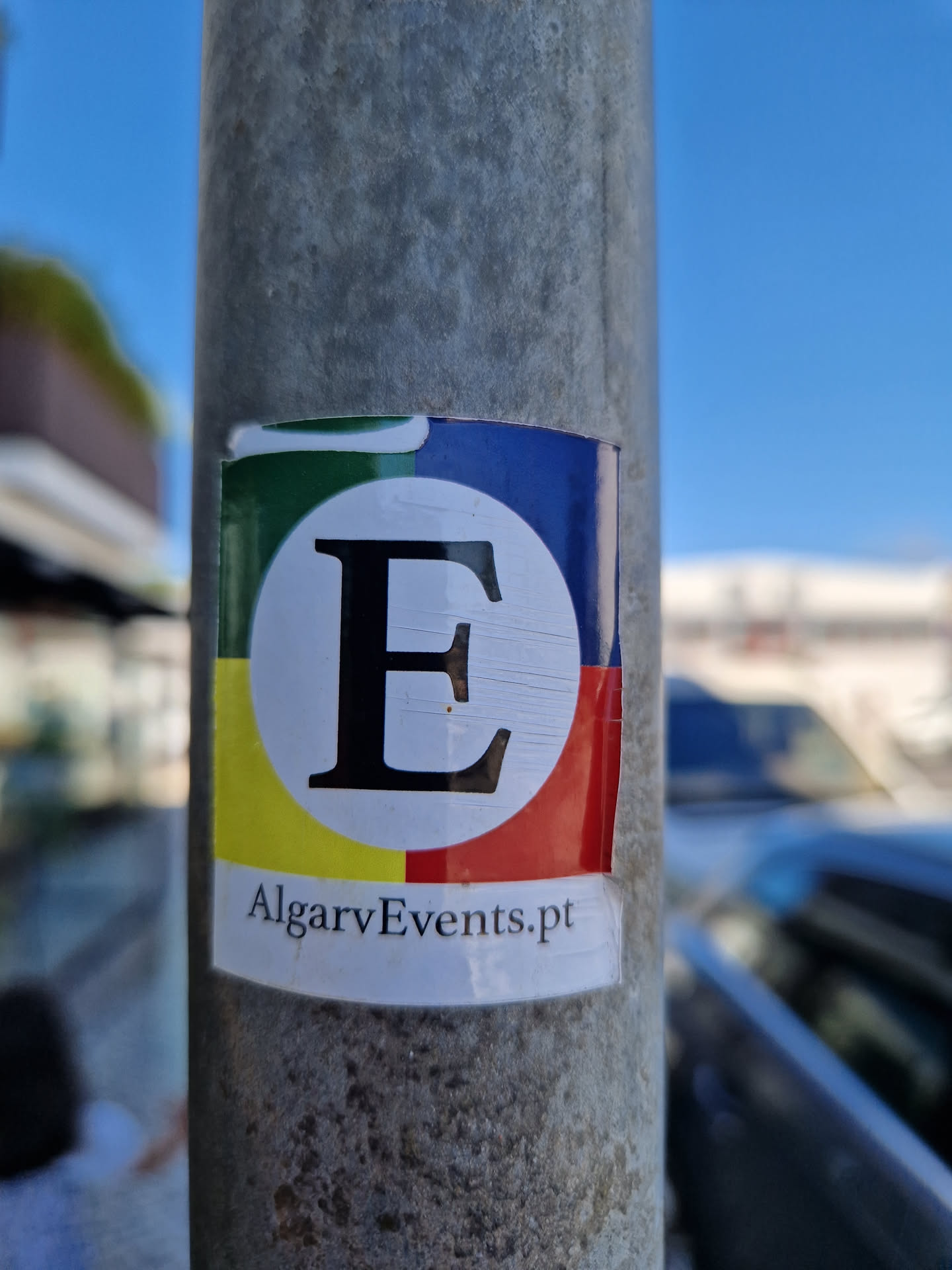 A sticker featuring a large letter 'E' in a circle with a color block design and the text 'AlgarveEvents.pt' below. The sticker is affixed to a metal pole.