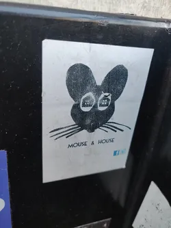 A sticker featuring a stylized illustration of a mouse's head. The mouse is depicted in black ink on a silver background. Below the illustration, the text "MOUSE & HOUSE" is visible, along with a small Facebook logo.