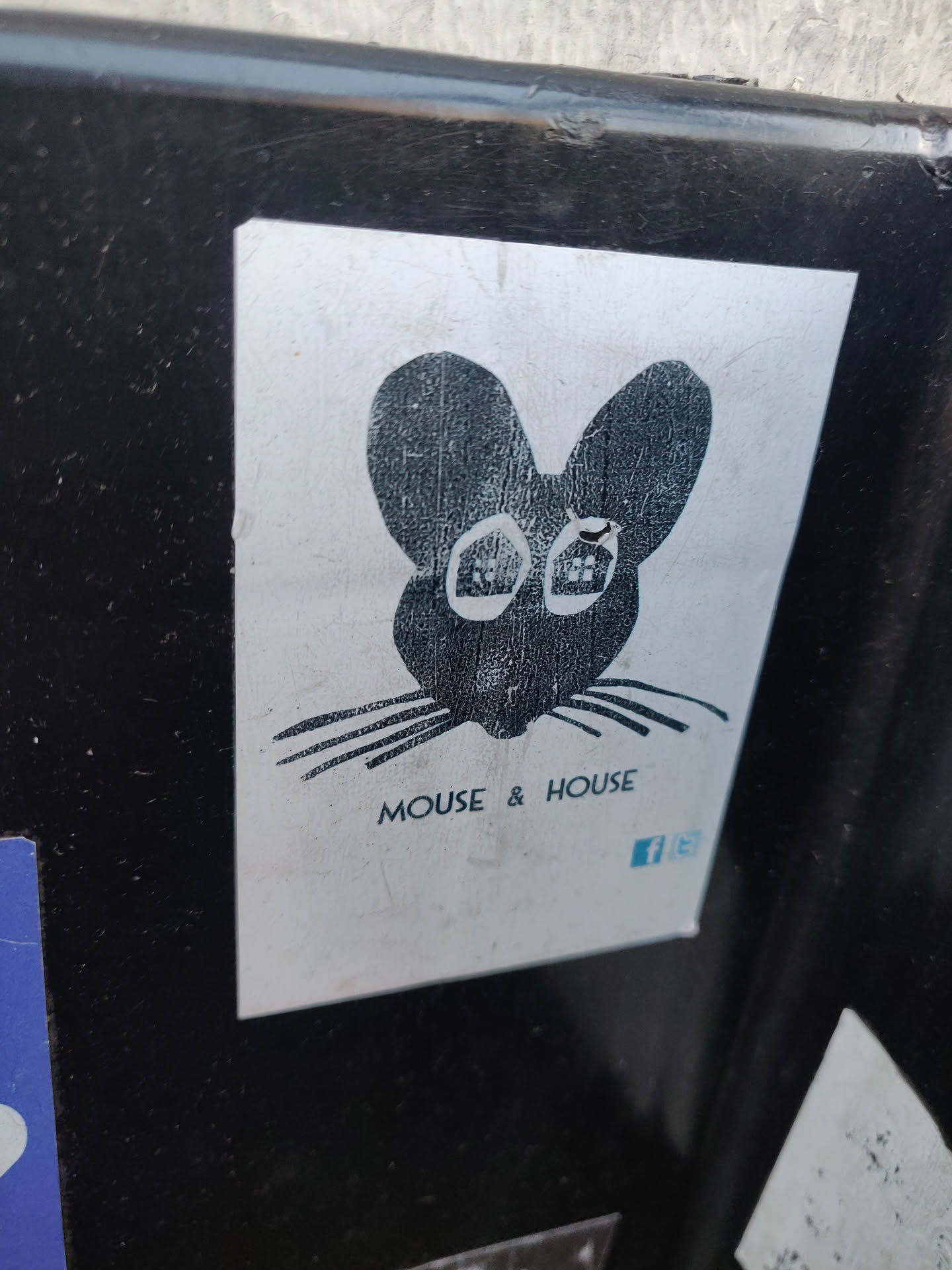 A sticker featuring a stylized illustration of a mouse's head. The mouse is depicted in black ink on a silver background. Below the illustration, the text "MOUSE & HOUSE" is visible, along with a small Facebook logo.