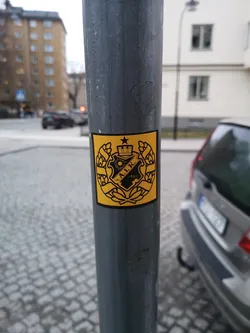 A square sticker with a vibrant yellow background and a detailed black design. The central element is a heraldic crest featuring a crown at the top, a five-pointed star to its left, and a stylized sun with rays. Within the shield of the crest, the initials 'A.I.K.' are prominently displayed, with the year '1891' beneath them. The entire crest is framed by two symmetrical laurel wreaths, signifying victory or honor. The design is intricate and monochromatic, using only black lines and solid shapes against the yellow.