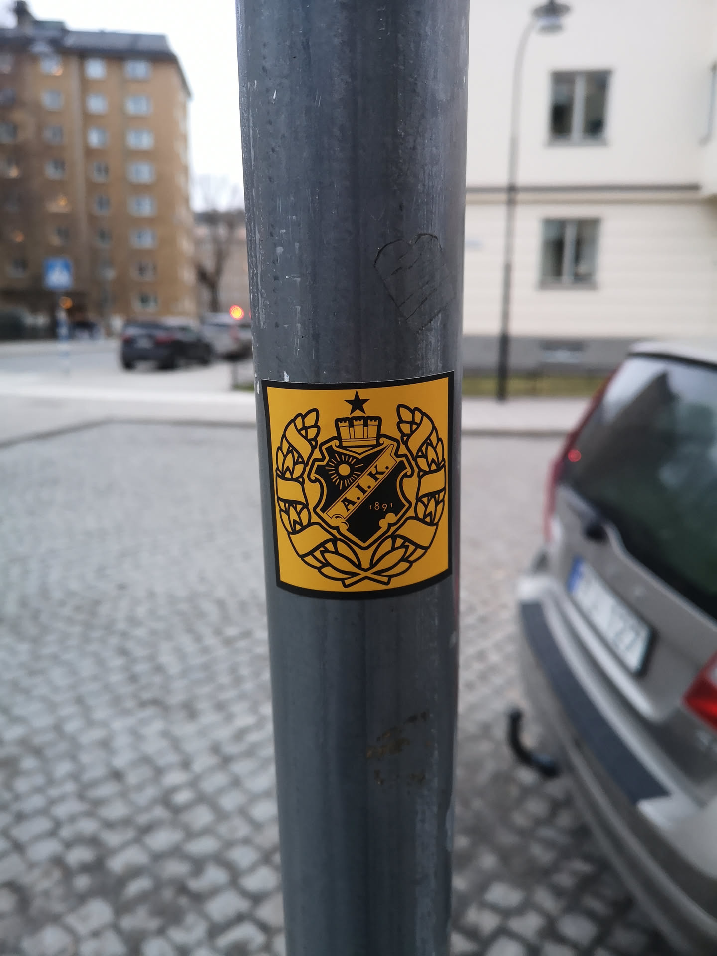 A square sticker with a vibrant yellow background and a detailed black design. The central element is a heraldic crest featuring a crown at the top, a five-pointed star to its left, and a stylized sun with rays. Within the shield of the crest, the initials 'A.I.K.' are prominently displayed, with the year '1891' beneath them. The entire crest is framed by two symmetrical laurel wreaths, signifying victory or honor. The design is intricate and monochromatic, using only black lines and solid shapes against the yellow.