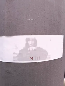 This sticker shows a person's photograph in grayscale with the text 'MTH' printed in reddish-brown font. The sticker is affixed to a gray, textured, cylindrical surface.