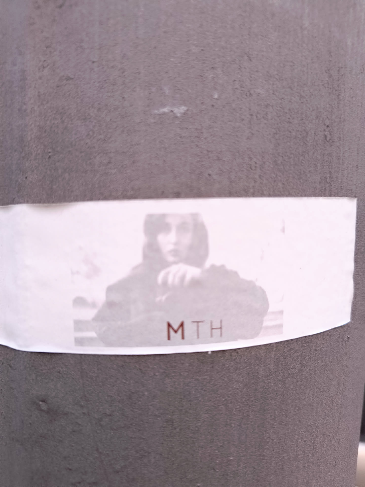This sticker shows a person's photograph in grayscale with the text 'MTH' printed in reddish-brown font. The sticker is affixed to a gray, textured, cylindrical surface.