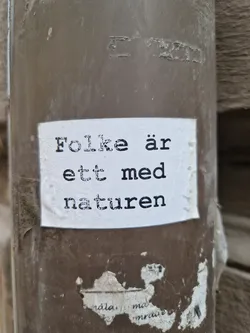 A sticker with the text "Folke är ett med naturen" (People are one with nature) in Swedish, attached to a brown pipe.