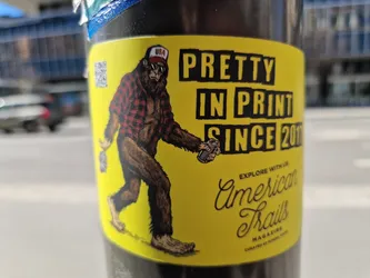 Yellow sticker featuring a bigfoot-like creature wearing a red and black plaid shirt and a baseball cap. The creature is holding a can and walking. The text on the sticker says "Pretty in print since 2016" and "American Trails Magazine".