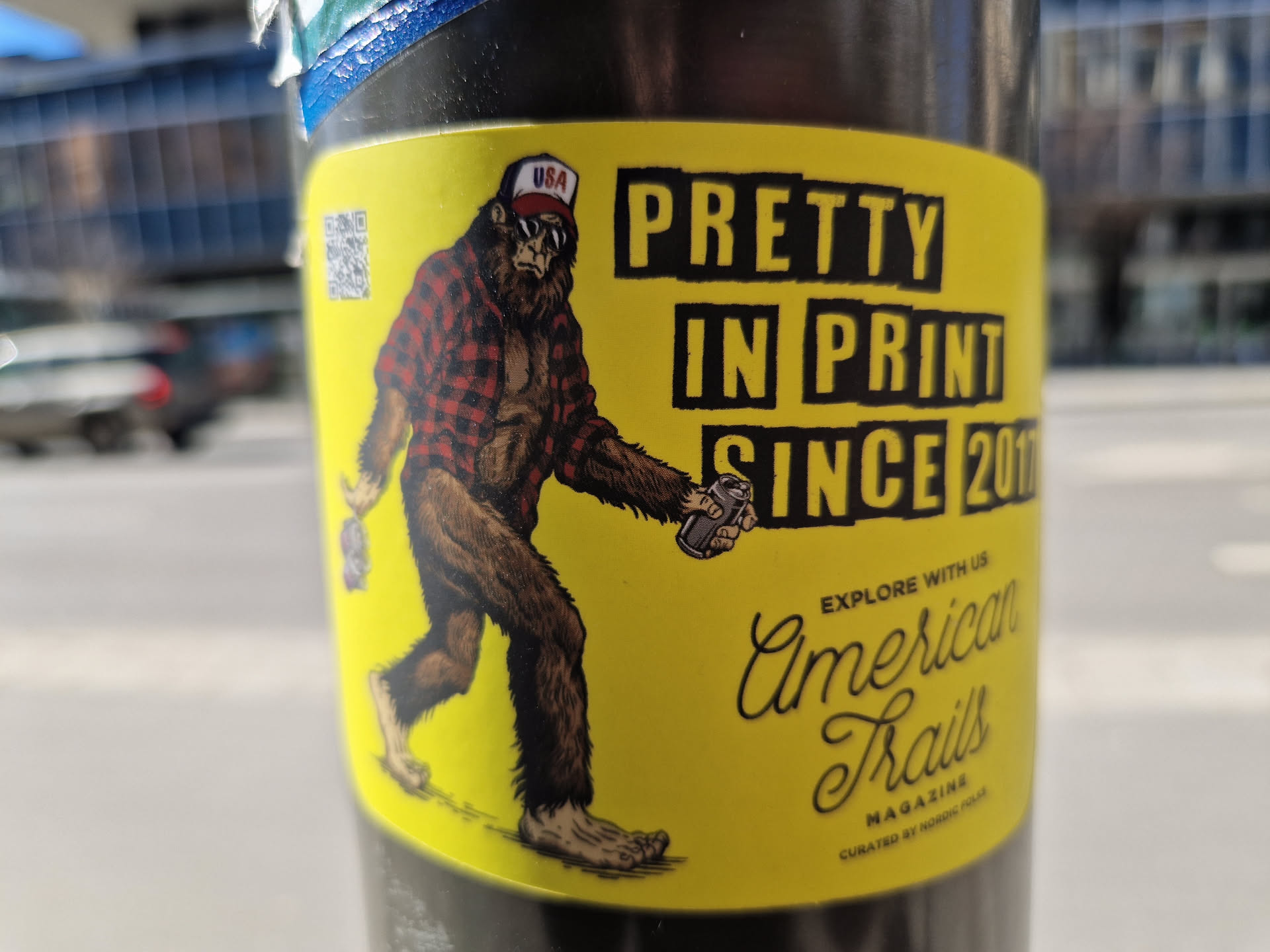 Yellow sticker featuring a bigfoot-like creature wearing a red and black plaid shirt and a baseball cap. The creature is holding a can and walking. The text on the sticker says "Pretty in print since 2016" and "American Trails Magazine".