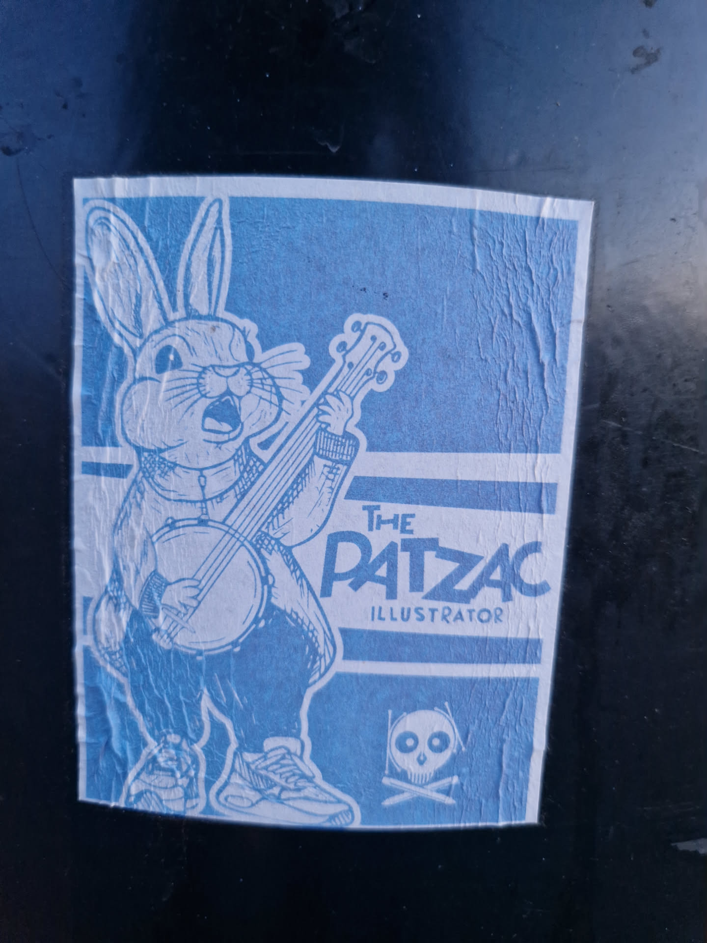 Blue sticker featuring an illustration of a rabbit playing a banjo. The text 'The Patzac Illustrator' is visible at the bottom. There is also a small skull and crossbones symbol.